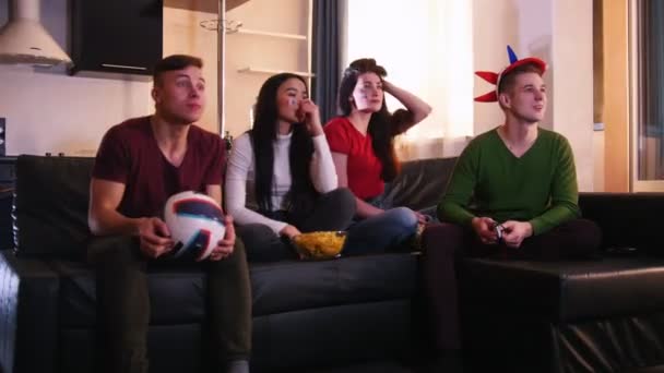 Czterech młodych ludzi siedzi na kanapie i oglądać mecz piłki nożnej. Zespołu trafi cel, jedna para są szczęśliwi i inny jeden rozczarowany — Wideo stockowe