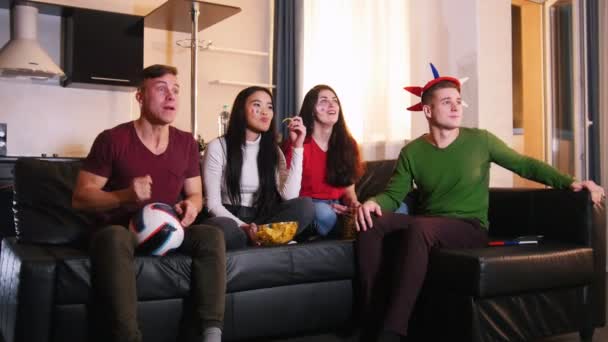 Fyra unga vänner sitter i soffan och tittar på en fotbollsmatch. Ett team träffar målet, ena paret är glada och en annan besviken — Stockvideo