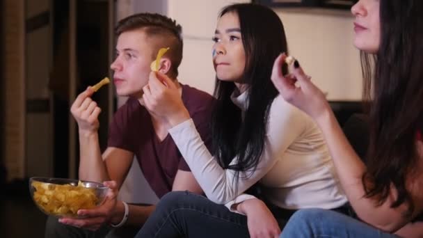 Четверо молодых людей смотрят футбольный матч, едят нездоровую пищу и разговаривают за игрой — стоковое видео