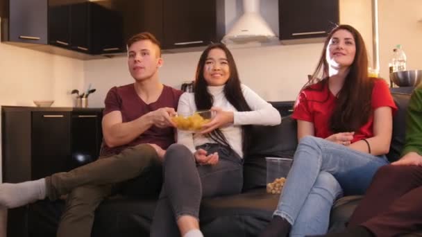 Companhia de jovens amigos que passam tempo juntos. Assistir TV e entregar uma tigela cheia de batatas fritas — Vídeo de Stock
