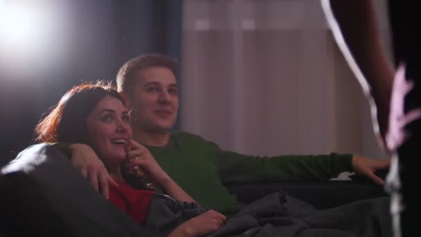 Giovane coppia sdraiata sul divano e guardando la TV, il loro amico arriva, si siede vicino a loro e stanno ridendo — Video Stock