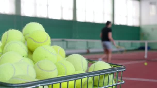 Два юноши играют в теннис на теннисном корте. Тележка с теннисными мячами на переднем плане — стоковое видео