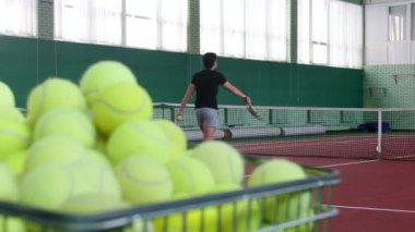 Bir sepeti tenis topları ile dolu. İki genç adam arka plan tenis kortunda tenis oynamak