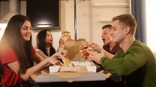 Company of young friends sitting in kitchen and eating pizza