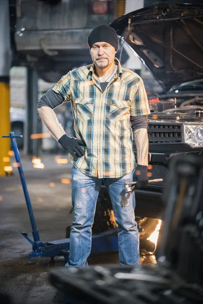 Usługi naprawy samochodów. Brutalny mechanik mężczyzna w czarnym kapeluszu stoi obok samochodu i trzymając klucz — Zdjęcie stockowe