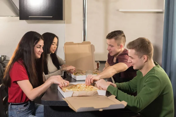 Company of four young friends sitting in kitchen and opening a pizza box