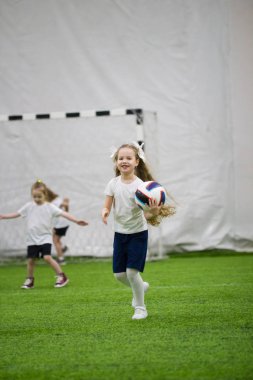 Çocuklar kapalı futbol oynuyor. Bir topu tutarak çalışan küçük bir kız