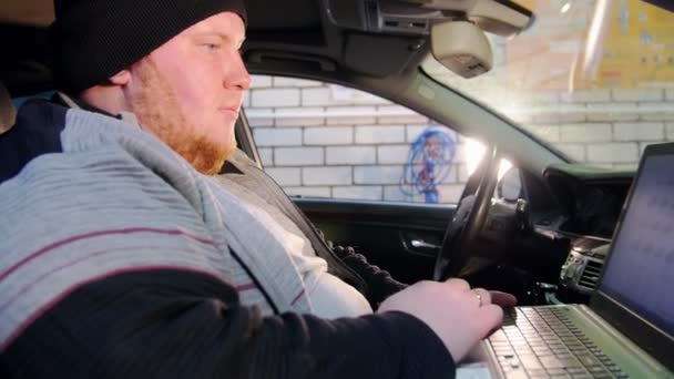 Servicio de coches. El hombre mecánico grueso se sienta en el coche y trabaja con un ordenador portátil — Vídeo de stock