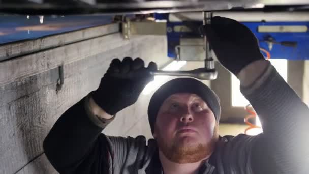 Автосервис. Толстый механик, стоящий в смотровой яме под машиной и работающий с инструментом — стоковое видео