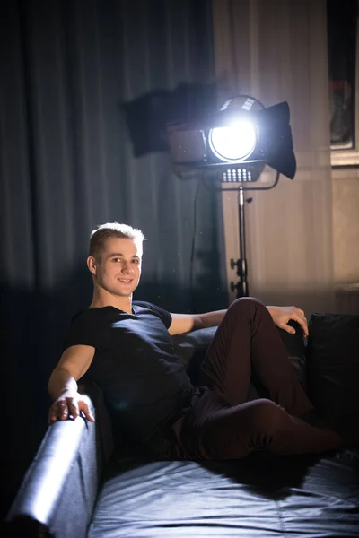 Ung attraktiv leende kille i mörk t-shirt som sitter på en soffa och tittar i kameran. Poserar. Bright belysning — Stockfoto