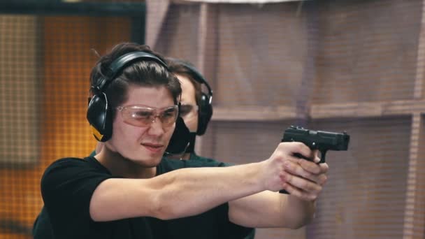 В тир. Концентрированный молодой человек в защитных очках и наушниках целится в цель и стреляет — стоковое видео