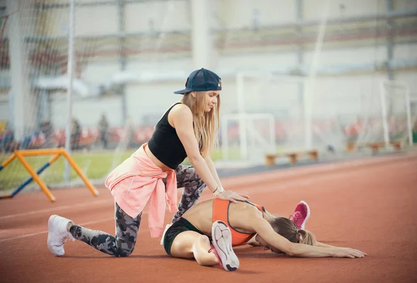 Young beautiful woman with blonde hair helps another woman to stretch her legs in the sports hall after training — Stock Photo, Image