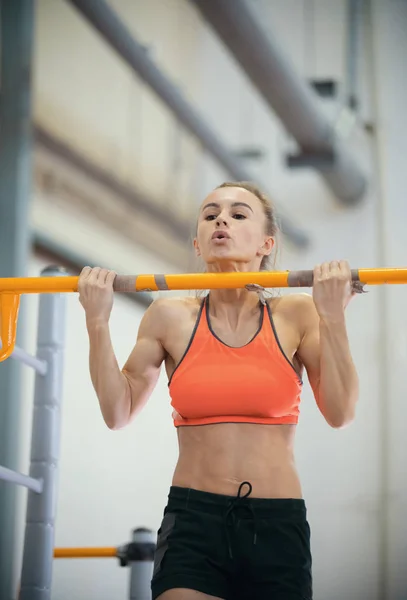 Mujer joven con el pelo rubio tira hacia arriba en el travesaño y la valentía duro en la sala de deportes —  Fotos de Stock