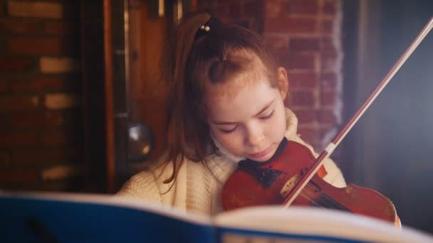 Музыкальный урок. Маленькая красивая девочка играет на скрипке по нотам — стоковое видео