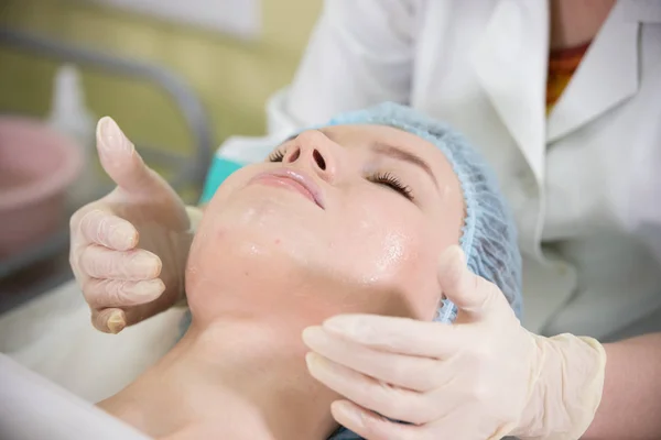 Klinik Kosmetologi. Tangan dokter melakukan pijat wajah untuk seorang wanita muda dengan mata tertutup berbaring di tempat tidur — Stok Foto