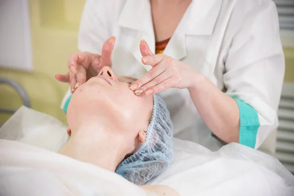 Primer plano de hacer un masaje facial a una mujer joven con los ojos cerrados en la clínica, médicos manos tocando las mejillas de los pacientes — Foto de Stock