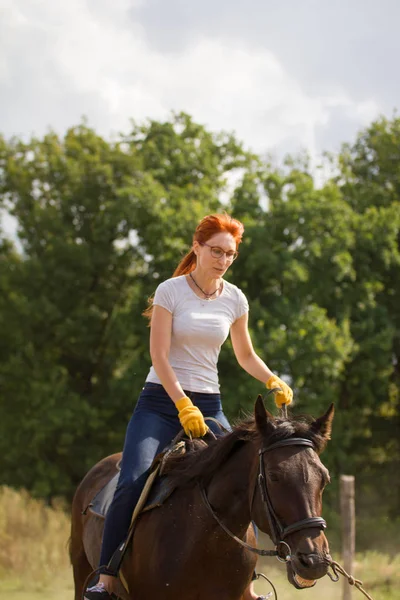 En rödhårig kvinna i vit t-shirt som rider en häst i naturen. — Stockfoto