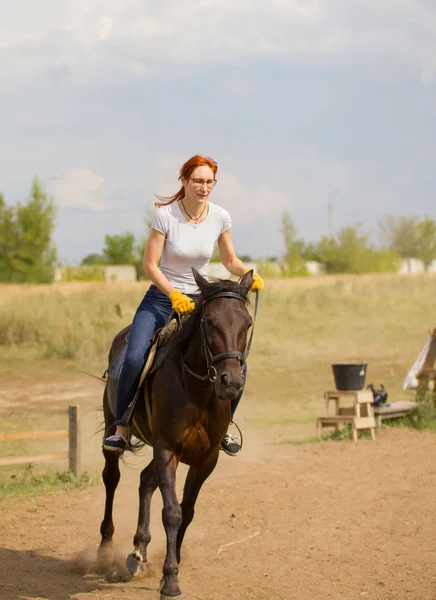 A redhead woman in yellow gloves riding a horse on the field. — Stock Photo, Image