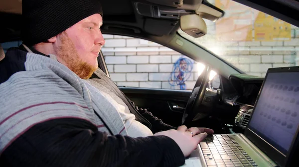 Servicio de coches. El hombre mecánico grueso se sienta en el coche y trabaja con un ordenador portátil — Foto de Stock