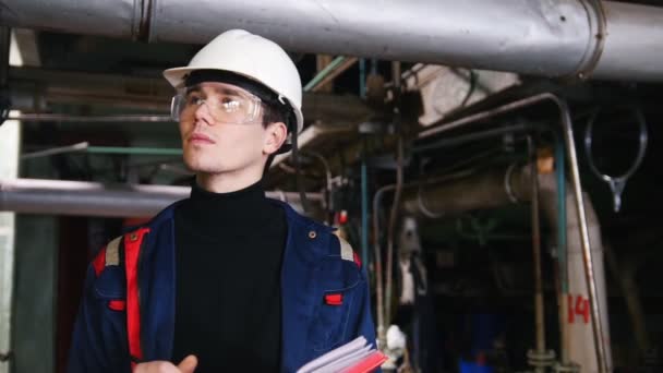 Un ingeniero hombre en un casco y gafas de protección haciendo notas en la planta de fabricación — Vídeos de Stock