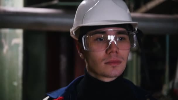 Un joven ingeniero en gafas protectoras caminando en planta de fabricación y mirando a su alrededor — Vídeos de Stock