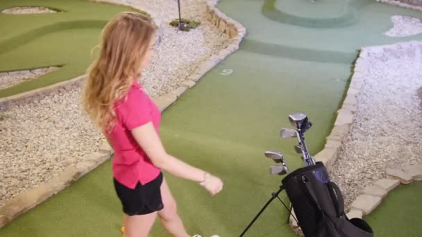 Gry w mini golfa. Młoda blond kobieta przychodzi do worka, biorąc kij golf i odchodzi — Wideo stockowe