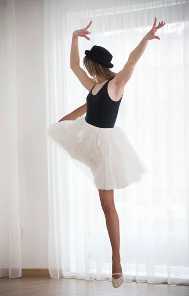 Ballerina in hat stands near the window, raises her leg, in a bright studio