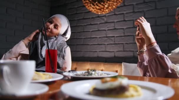 Две мусульманки сидят за столом в ресторане. Женщина, фотографирующая своего друга — стоковое видео