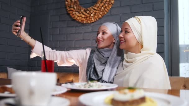 Δυο μουσουλμάνες που κάθεται σε ένα εστιατόριο και λαμβάνοντας μια selfie στο τηλέφωνο — Αρχείο Βίντεο