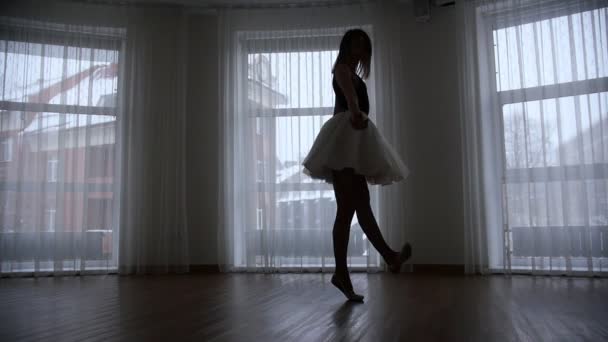 Ένα στούντιο στο λυκόφως. Μια σιλουέτα της νεαρής γυναίκας μπαλαρίνα εκτελεί μια περιστροφή μπροστά από το παράθυρο — Αρχείο Βίντεο
