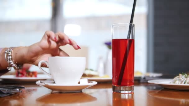 Una taza de café y un vaso de bebida sobre la mesa. Una mano de mujer toma el azúcar y lo pone en la taza, otra mujer toca la paja — Vídeo de stock