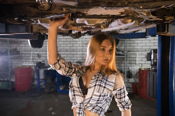 Joven mujer mecánica sexy en camisa desabotonada a rayas de pie debajo del coche en el servicio de reparación de automóviles — Foto de Stock