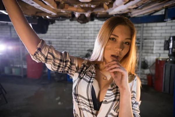 Ung sexig mekaniker kvinna i uppknäppt skjorta stående under bilen i bil reparationsservice. Titta i kameran — Stockfoto