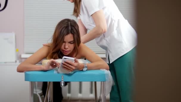 Een jonge vrouw ontvangt een ontspannende rug massage. Vergadering in haar telefoon in sociaal netwerk — Stockvideo