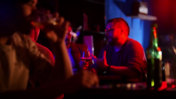 Bar con illuminazione al neon. Compagnia di amici che bevono birra. Un giovane uomo in primo piano che beve birra — Video Stock