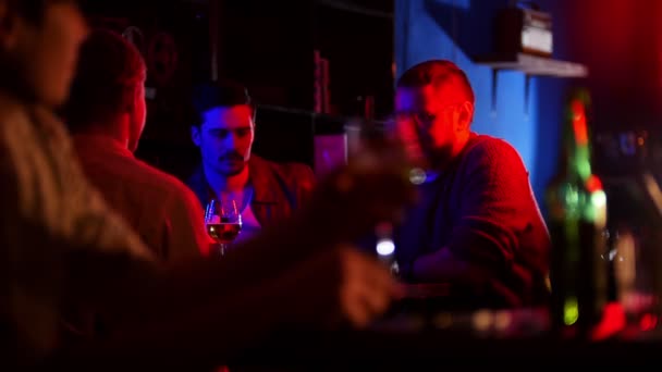 Bar com iluminação de néon. Companhia de amigos a beber cerveja. Um jovem em primeiro plano vê seus amigos e se senta ao lado deles — Vídeo de Stock