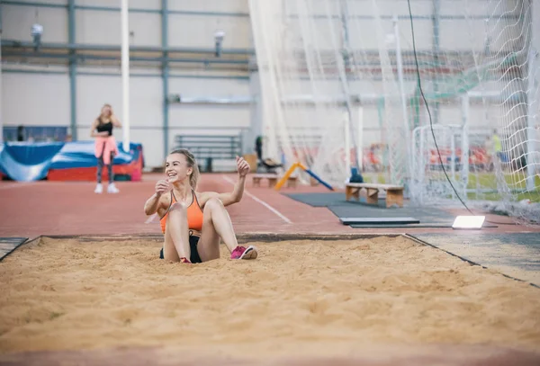 Νεαρά ξανθιά, χαμογελώντας γυναίκες εκτελούν ένα μεγάλο άλμα στον αθλητικό στίβο. Κάθεται στην άμμο — Φωτογραφία Αρχείου
