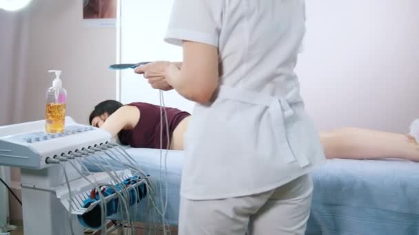 Clínica de Cosmetología. Mujer joven tomando mioestimulación, acostada en la cama. Un médico adjuntando un equipo — Vídeos de Stock