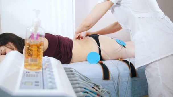 Kosmetika klinika. Mladá žena s myostimulation, ležící v posteli. Doktor, připojování zařízení k tělu — Stock video