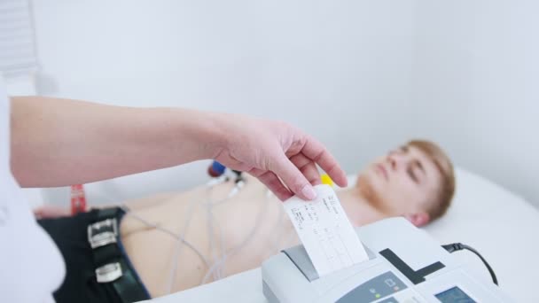 A medical clinic. A young man receiving a ECG procedure. A results tape coming out of machine — Stock Video