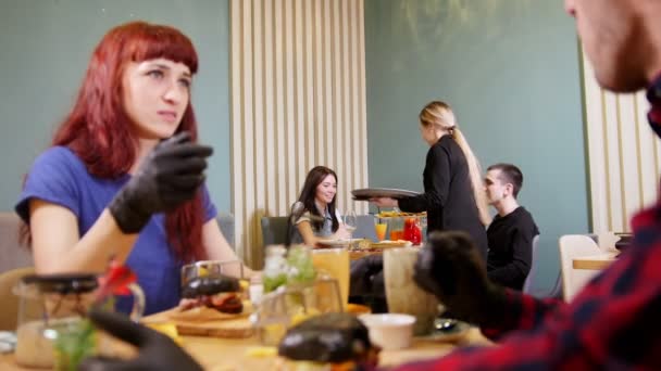 Två personer i svarta handskar med svart hamburgare och äta den. Att se sina vänner av en annan tabell och vinkar dem med en hand. En servitör leenden — Stockvideo