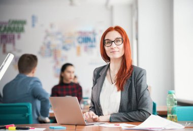 Zencefil gülümseyen kadın ofiste oturan ve laptop yanında çalışma gözlük. Kamerada arıyorsunuz