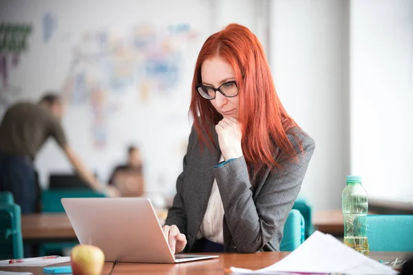 Рыжая задумчивая деловая женщина в очках, сидящая в офисе и работающая за ноутбуком. Глядя на экран — стоковое фото