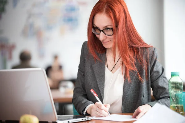 Рыжая деловая женщина в очках сидит в офисе, смотрит на ноутбук и делает заметки — стоковое фото