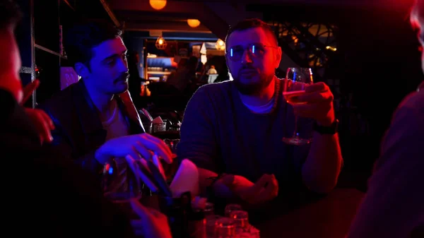 Bar com iluminação de néon. Grupo de amigos sentados à mesa e bebendo álcool enquanto conversam — Fotografia de Stock