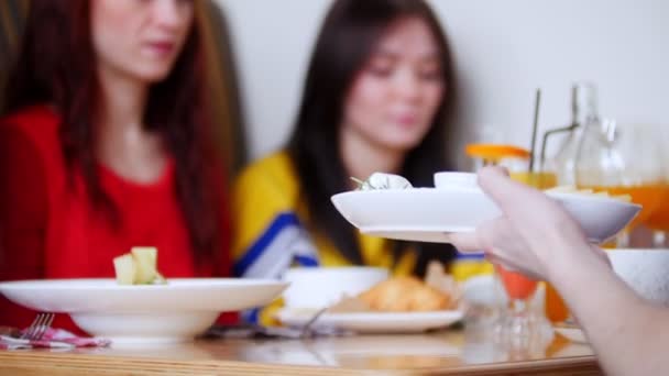 Mujeres amigas sentadas en un café. Mesa llena de diferentes alimentos y bebidas. Una persona frente a ellos toma el plato — Vídeos de Stock