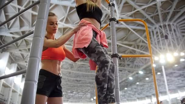 Fiatal nő húzza fel a keresztléc a sportcsarnokban. Szivattyú alsó része a testét, tréner, segít neki — Stock videók