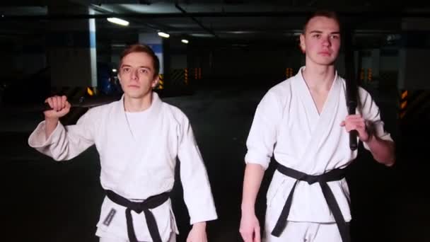 İki genç adamın omuzlarında bir eğitim kılıç tutan bir otopark yürürken kimono içinde — Stok video