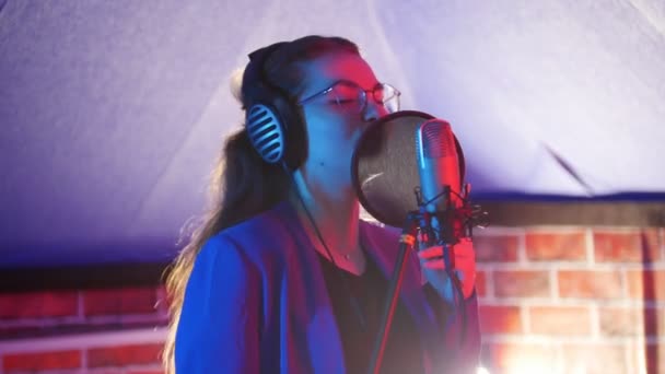 Uma jovem mulher em óculos e fones de ouvido cantando no estúdio — Vídeo de Stock
