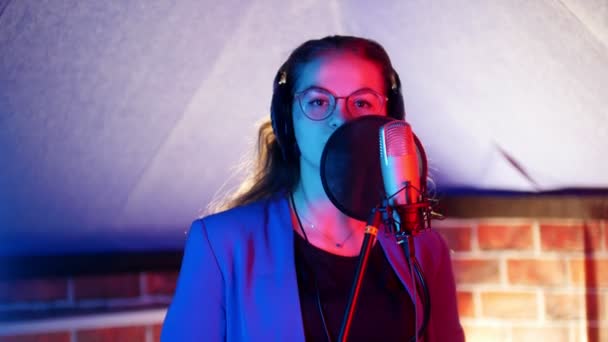 Una giovane donna con occhiali e cuffie che canta in studio. Guardando nella macchina fotografica — Video Stock